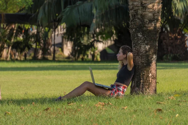 young pretty student in black top with laptop on knees sits under palm tree