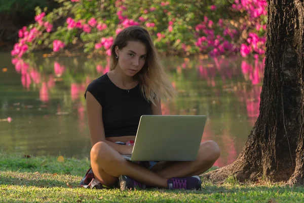 nice fair-haired girl working on her laptop near pond sitting in lotus pose,