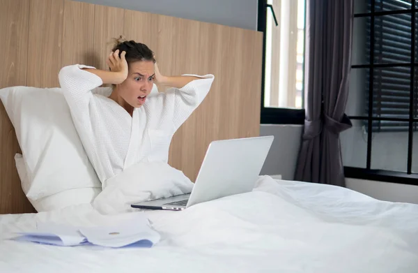 blonde in bathrobe sits in her bedroom, pillow behind her back, laptop on legs