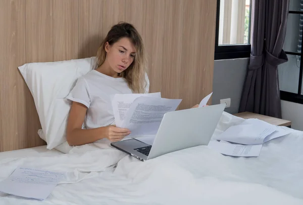 Young student working at home in her bedroom, reading the papers, sitting in bed