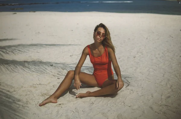 nice shot of fit model in orange swimsuit siting on the white sand in the shade