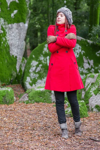 Young woman lost on a forest shivering with cold and embracing or holding herself, wearing a red long coat or overcoat, a beanie and gloves during fall, autumn or winter