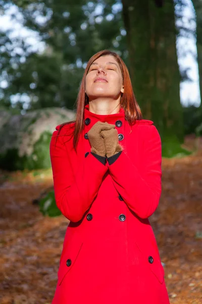赤いロングコートを着て祈りと冬の日光の暖かさを楽しむ森の女性 — ストック写真