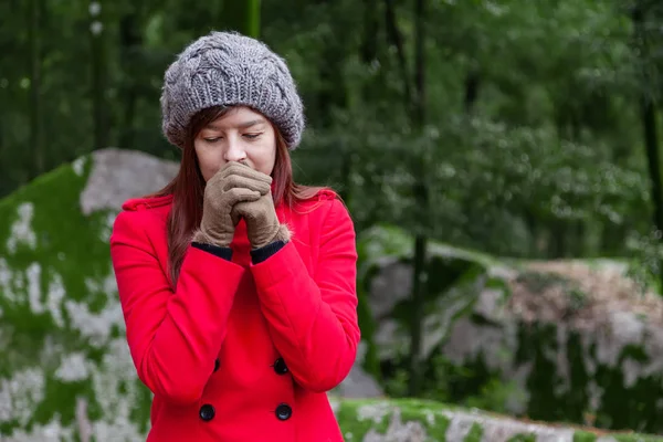 Young woman shivering with cold on a forest wearing a red overcoat, a beanie and gloves during winter