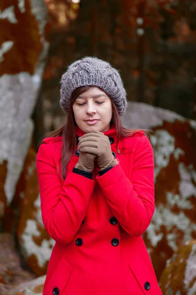 Young woman shivering with cold on a forest wearing a red overcoat, a beanie and gloves during winter