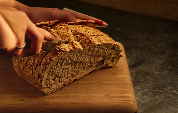 45 degrees picture of natural bread with seeds, over wood table, black background, and hand with knife cutting a slide of bread, including red and brown cloth