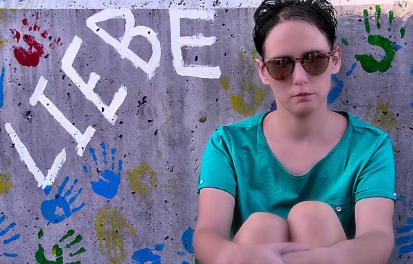 Straight on photo of sat woman, concrete background with words and painted children hands, looking at camera