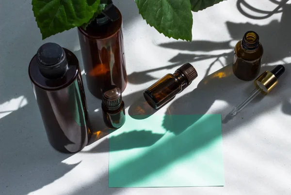 Dark cosmetic bottles and green natural leaves on a light background. Green empty card, sheet for writing. Layout for adding inscriptions. Natural hard light, deep shadows. The concept of natural envi