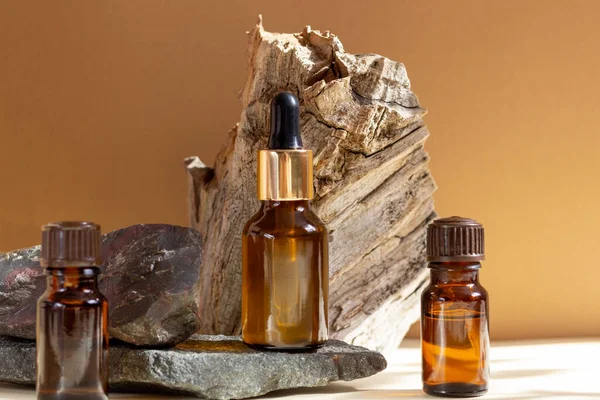 Glass cosmetic bottles with a dropper stand next to a log on a beige background with bright sunlight. The concept of natural cosmetics, natural essential oil.