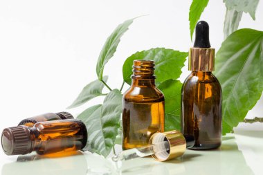 Beyaz arka planda yeşil yaprakların yanında damlaları olan cam kozmetik şişeler. Organik kozmetik konsepti, doğal temel yağ ve krem.