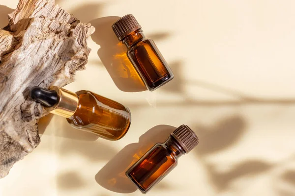 Glas cosmetische flessen met een druppelaar staan naast een log op een beige achtergrond met fel zonlicht. Het concept van natuurlijke cosmetica, natuurlijke essentiële olie — Stockfoto