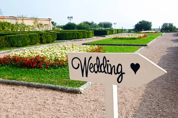 Wedding sign made with stylish black handwritten font and the shape of heart on white wooden arrow in beautiful park with green lawns and cut bushes