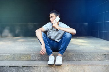 Genç bir çocuk cep telefonuyla dışarıda sohbet ediyor. Şehir merdivenlerinde oturuyor. Başını yana çevirip düşünceli bir ifadeyle sohbeti dinliyor.