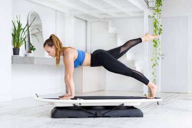 Formda sörf tahtası yapan atletik formda genç bir kadın yoga pozu veriyor. Yüksek anahtarlı spor salonunun yan görüntüsünde, sağlık ve zindelik konseptinde taze yeşil bitkilerle birlikte.
