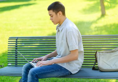 Parkta dizüstü bilgisayarını kullanan genç bir çocuk güneşli bir yaz gününde yeşil çimen arka planına karşı bankta oturmuş daktiloyla onu dengeliyor.