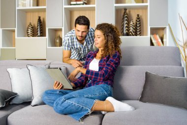 Genç bir çift, kişisel bir eğlence konseptiyle evlerinin oturma odasında rahatlarken birlikte tablet üzerinde medyayı seyrediyor.