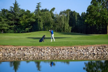 Golfçü su kenarında golf sahasında güzel bir atış yapıyor, suya yansıyor.