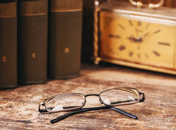 The glasses lie on a wooden shelf on the background of books and the old watch — Stock Photo, Image