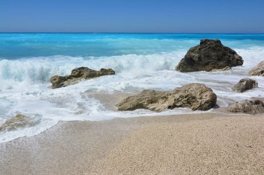 Kathisma sahilinin mavi ve beyaz dalgaları koyu kayaların üzerinde Kathisma sahilinin sığ sularında, Lefkada, Yunanistan