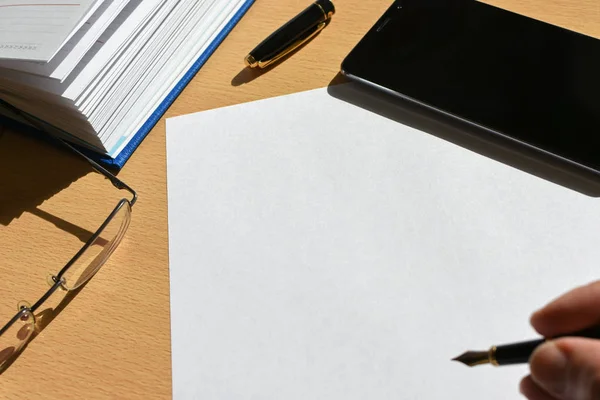 Hand writing pen on a sheet of paper on a wooden desktop in the office, smartphone, stationery, glasses, copy space — Stock Photo, Image