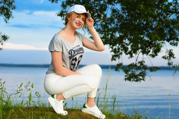 Romantic young girl in sports clothes with a cap in the sunlight against the backdrop of the shore of the lake water in the evening — Stock Photo, Image