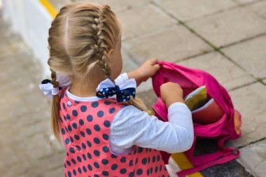 Küçük kız dergi, kitap ve aksesuarları bir sırt çantası içinde bir okul bahçesinde koyar