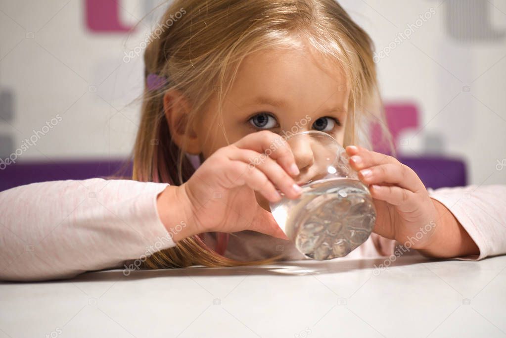 Little girl drinks water from glass at the table