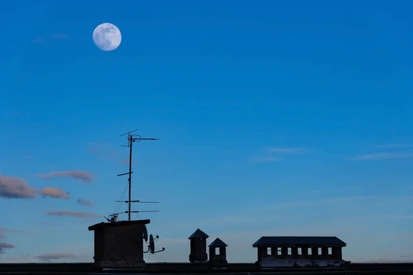 Silhouettes d'antennes et conduits de ventilation sur le toit de la maison contre le ciel bleu du soir avec la lune — Photo