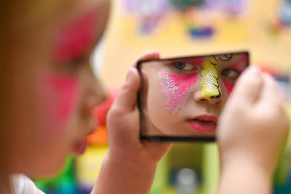 A little girl with a painted face looks at herself in the mirror at the party