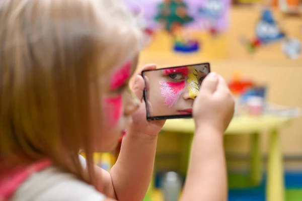 Une petite fille au visage peint se regarde dans le miroir à la fête — Photo