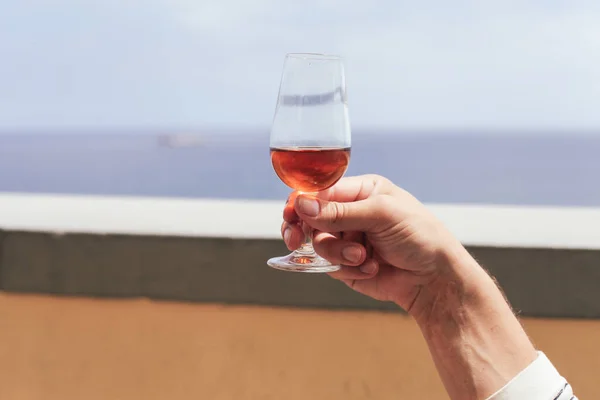 Elinde bir bardak Madeira şarabı, arkasında mavi deniz.
