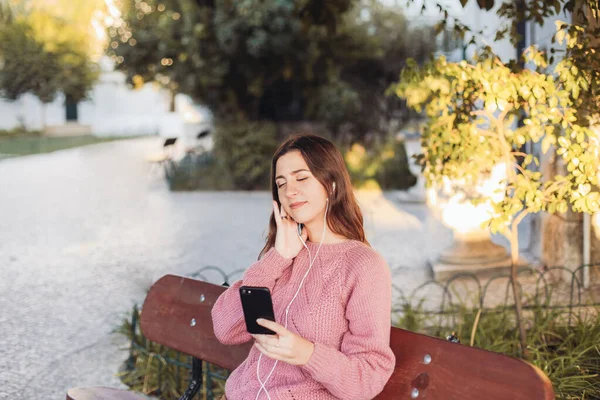 Kulaklıklı genç bir kadın bankta oturuyor ve akıllı telefonundan müzik dinliyor, gözleri kapalı. Arka planda sonbahar parkı 