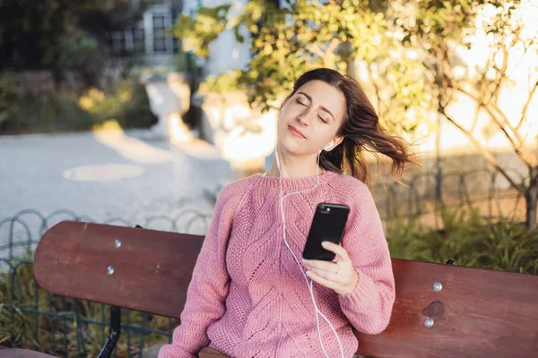 Kulaklıklı genç bir kadın bankta oturuyor ve akıllı telefonundan müzik dinliyor, gözleri kapalı. Arka planda sonbahar parkı 