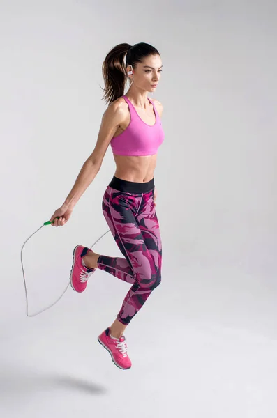 Sporty beautiful young woman with jump rope makes fitness exercising at gray background. Copy space