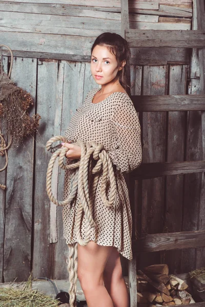 Lovely girl in a modest dress with long solid piece in rope in her hands standing in a planked barn. Looking at camera