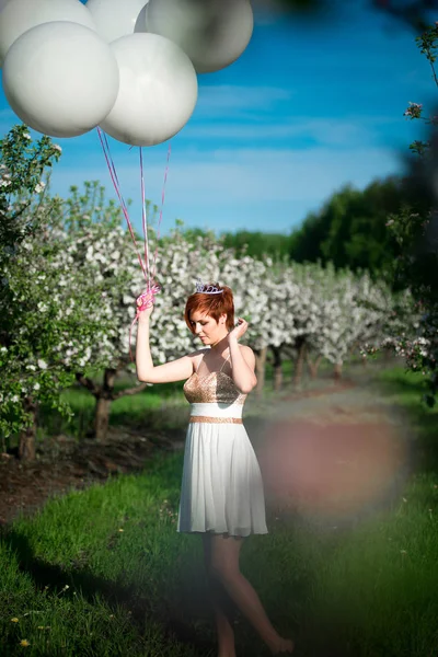 Charming girl in the green garden with balloons tied to her hand fixing her red short hair