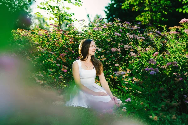 Dark haired girl with a dreaming look in a white dress sitting in flower bushes