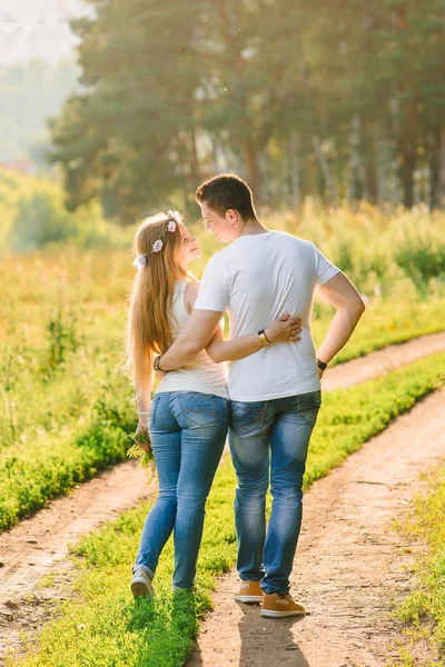 Couple in blue jeans and white t-shirts looking at each other happily standing back to the camera green grass and trees all around their path