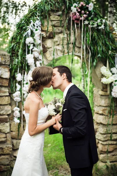 Nygifte Kysser Bryllupsseremonien Utenfor Grønt Område Steinbuen Med Blomster Ryggen – stockfoto