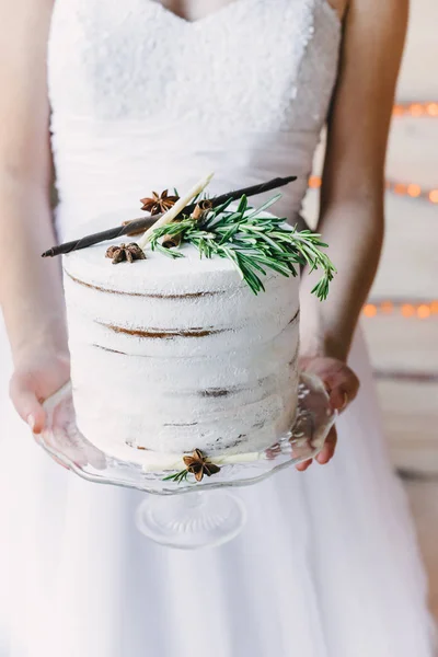 Bride holding a craft cake on a glass stand, wedding dress on the background