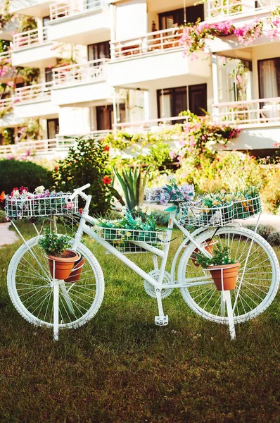 Decorative white bike with flower pots, nice hotel on the background. The white bicycle in the green garden
