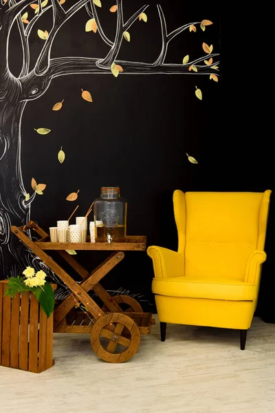 明亮的黄色扶手椅旁边的木制马车与纸眼镜和一个大瓶子上 黑墙与秋季画的背景 — 图库照片