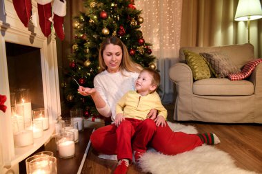 Genç çocuk annesi dizlerinin üzerinde oturan, birlikte mutlu görünüyordu yasaktır eşleştirme tutuyor. Noel oda iç tasarım, Xmas ağacı ışıkları tarafından dekore edilmiş