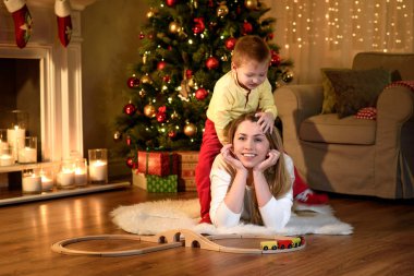 Sevimli küçük oğlan yalancı annesinin geri tırmanma, yeni yıl geleneksel olarak oda dekore edilmiştir. Mutlu Noeller ve mutlu yeni yıl. Güzel genç anne ile oğlu