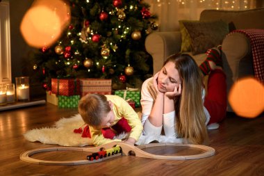 Genç Anne oğlunu merakla bir oyuncak tren ile oynama izliyor. Toddler anne Noel ağacı ile festively bir şekilde dekore edilmiş oda ile. Anne ve bebek çocuk portresi