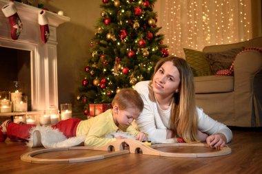 Şirin anne oğlu bir şirin ahşap tren ile oynamaya yakın fotoğraf için gülümsüyor. Genç Anne ve oğlu Noel arifesinde bir rahat karanlık oturma odasında şömine başına oturmuş