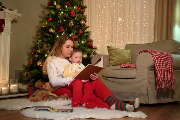 Mother is reading a fairytale from a big book to her son, he is hugging a toy rabbit, christmas atmosphere in a room. Happy family reading together on Christmas evening. Happy family New Year concept