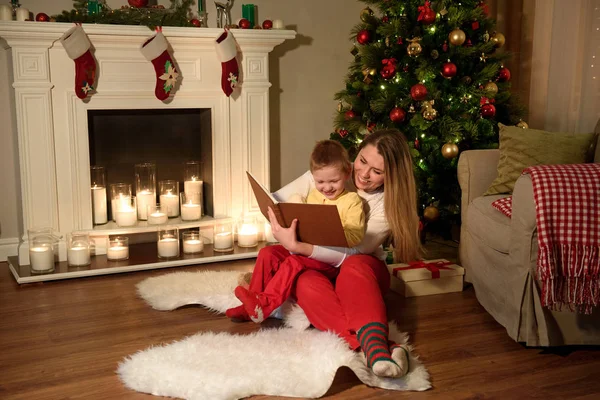 Mom and her boy are laughing on something from the book they are reading, holiday decorations in their house. Family with child celebrating Xmas. Decorated living room with tree,fire place and candles