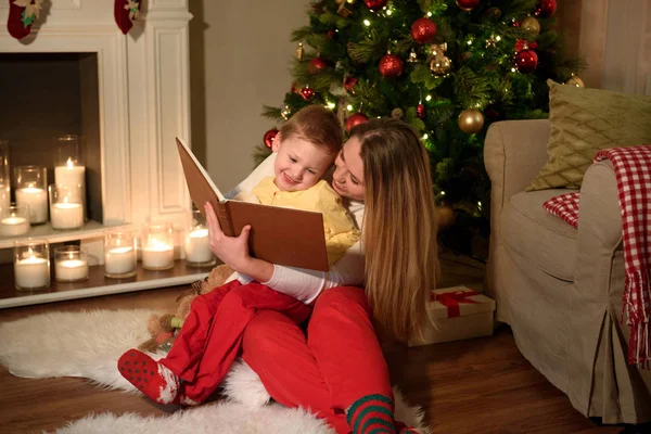 Bir çocuk ve onun anne ailesi bir güzel kitap gülüyor birlikte, rahatça Noel oda dekore okuyor. Mutlu bir aile birlikte Noel akşamı okuma. Kış akşam evde ebeveynler ve çocuklar için