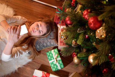 Güzel bir kadının telefonunu Noel tarafından çevrili ellerini tutarak üstten görünüm sunar. Kız köknar ağacı dalı ile yerde yatıyor. Genç kadın metin mesajlarını cep telefonu üzerinde kontrol